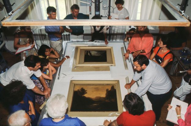 Bird's-eye view photograph of a group of people examining two paintings atop a table.