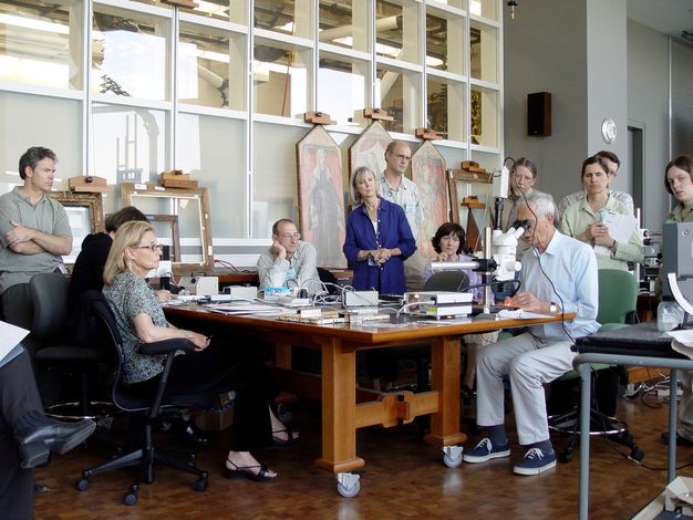 A group of people are centered around a man in a conservation lab, watching him work with his hands under a microscope.