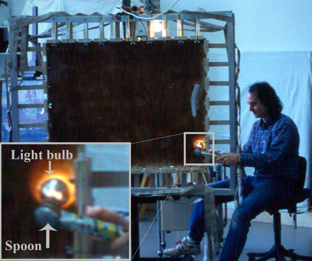 A man sits in a conservation lab holding a spoon tool up to a large painting on canvas with a lightbulb shining on the reverse side of the canvas. There is a detail showing the spoon and lightbulb being pressed at the same point on either side of the canvas.