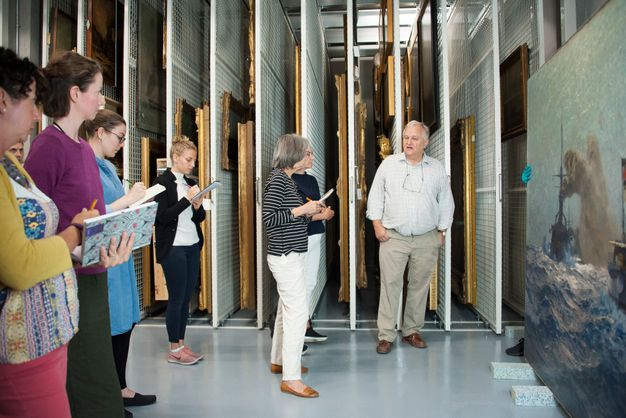 Photograph of people standing (left) while observing a seascape painting (right). A man stands and speaks in front of the painting.