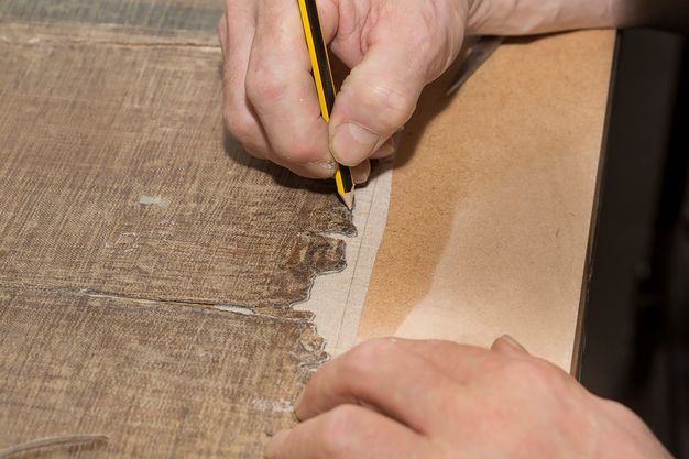 Photograph close-up of hands atop a table. The right hand holds a pencil and traces the outline of a frayed canvas.
