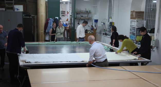 Photograph of people standing around a large table. An encased painting sits atop the table as a few people examine it.