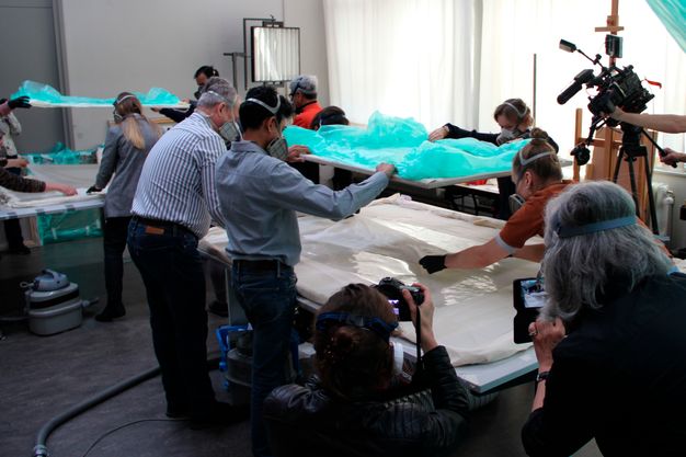 Photograph of people standing around painting that lays flat on a table. They are placing a blue-green cloth over painting.
