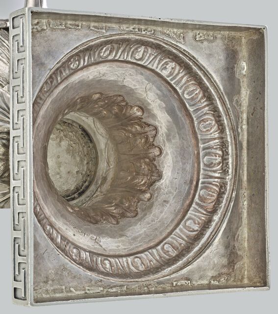 Close-up of a silver fountain's square base from an angle, revealing six rings of ornamentation making up the hollow base.