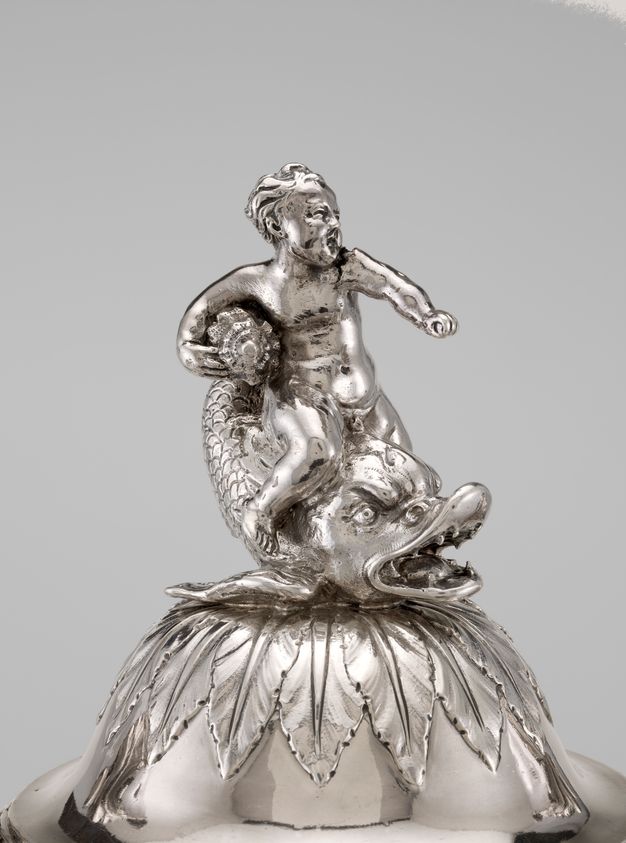 The lid of a silver vessel featuring a boy holding a conch shell sitting atop a dolphin and leaves.