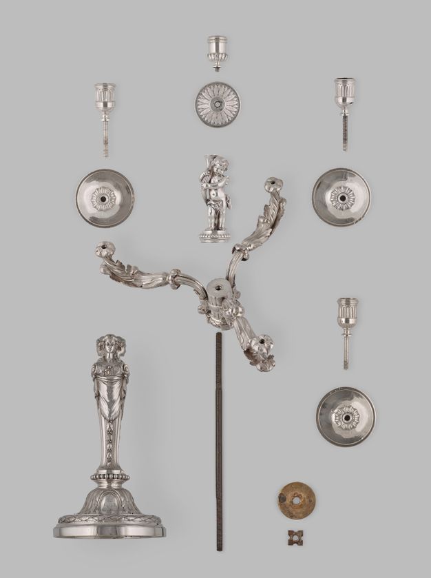Top view of the fourteen girandole parts, including four candle cups, four drip pans, a putto holding a branch, a head with three branches protruding from it, and the base of three women.