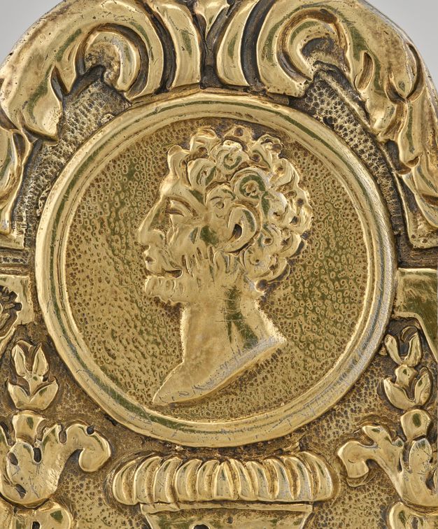 Profile of a man with curly hair and a beard framed by a circle and leafy decorations.