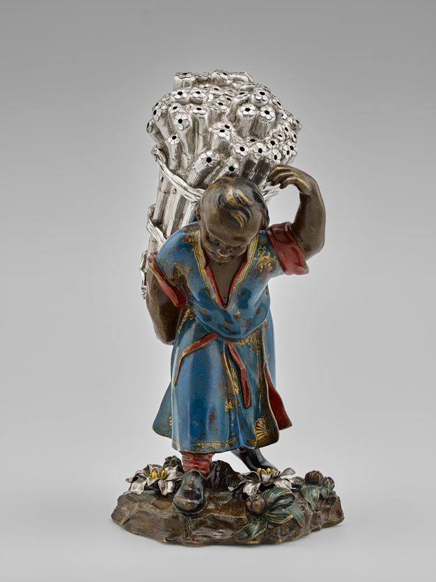 Front view of a field laborer carrying a bundle of sugarcane stalks atop a flower-covered mound.