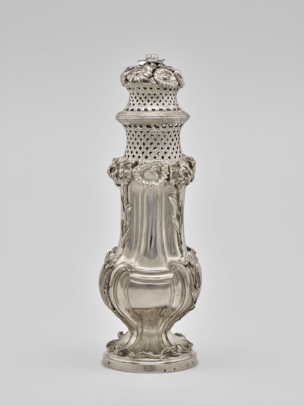 Silver sugar caster with a narrow leafy base, flowers rising up its body, and two woven baskets with flowers on top.