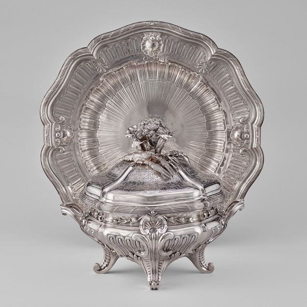 Silver lidded turren and liner topped with a cauliflower and lobster in front of a flower-shaped stand.