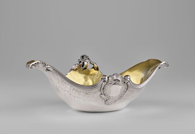 Angled view of the sauceboat with a high prow, low stern, lip rims that curl downward, and a shell-bordered oval surrounded by olive branches at the midpoint.