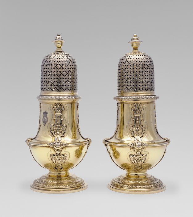 Pair of gold-colored sugar caster topped with an orb, cross-hatching pattern with holes, and a string of decorative flourishes down its center.