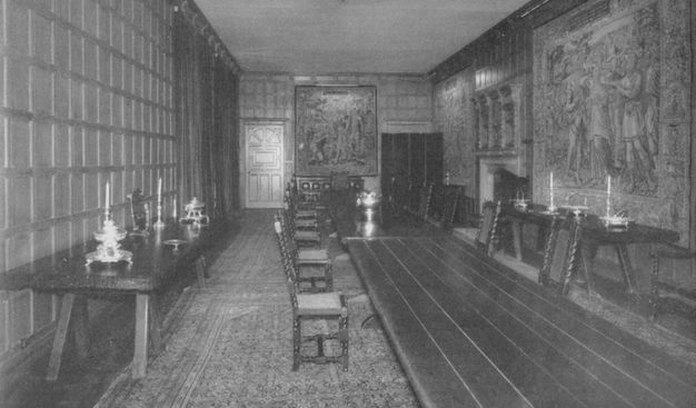 Dining room with a a pair of tureens on the side table at the left, a painting at the back wall, a tapestry on the right wall, and a long dining table running down the center.