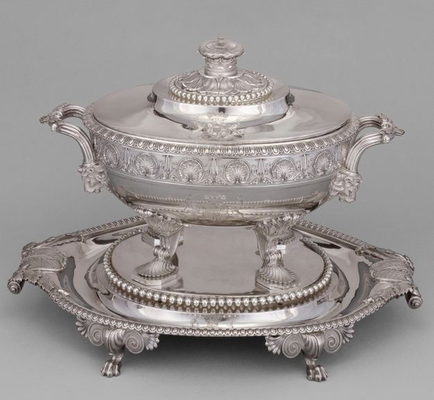 Silver tureen with shells decorating its body and shell-shaped legs standing on top of a raised stand with leafy accents.