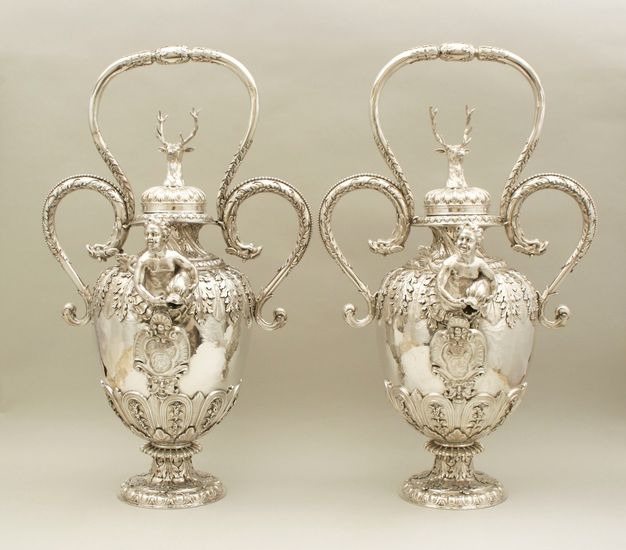 Two identical silver vessels with looping handles, decorated by a deer head with antlers on the lid, an adolescent holding open a dolphin's mouth coming out the center, and foliage wrapping the handles and base.