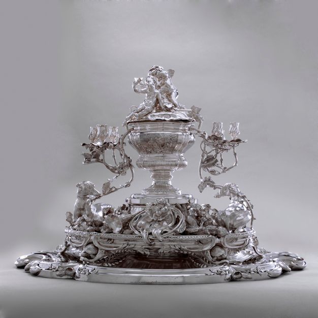 Stand and tiered tray set with greyhounds, furling foliage, a hunting horn, and dead game rising to support a coverned urn with three putti playing with stars on the lid. Two branches extend from the urn and transform into candelabra at its ends.