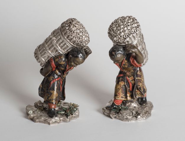 Two field laborers carrying sugarcane stalks on their backs wearing red, black, and gold robes atop of a base with yellow and green flowers.