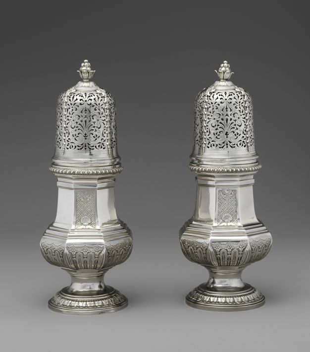 Two silver pear-shaped sugar casters with delicate flourishes on the lid and leafy patterns on the body.
