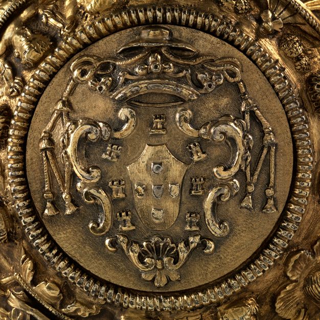 Close-up of a bishops's hat and prince's coronet on a coat of arms surrounded by tassels on either side on a gilded-silver salver on a stand.