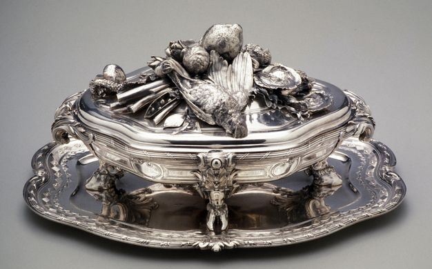 Silver lidded tureen topped by a dead bird, vegetables, and shellfish with four boar feet on top of a tiered tray.