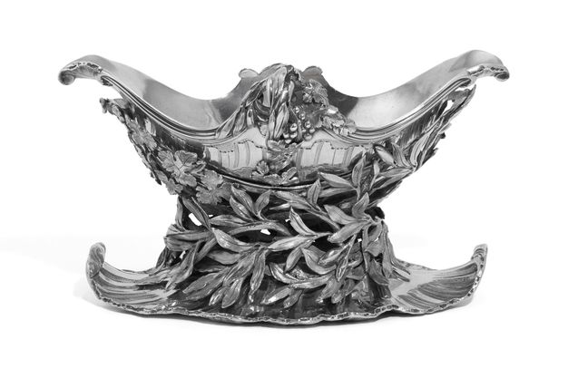 Silver sauceboat on a stand with grapes and leaves decorating its midpoint, a cradle covered in olive leaves, and a shell-edged base.
