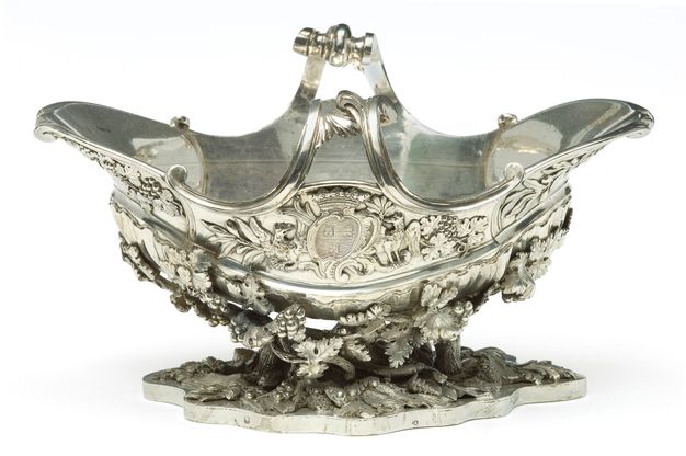 Side view of a sauceboat with a coat of arms at its midpoint and grape vines covered in fruit cradling the boat.