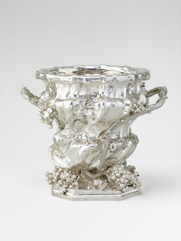 Tall silver vessel with handles disguised as grape vines and bunches of grapes decorating its body and covering its base.