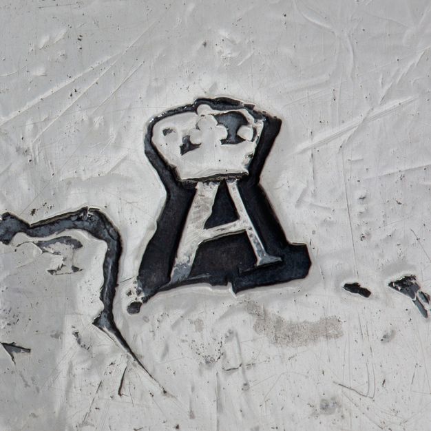 Close-up of a crowned A stamped on the stand, partially obstructed by another stamp on the bottom left.