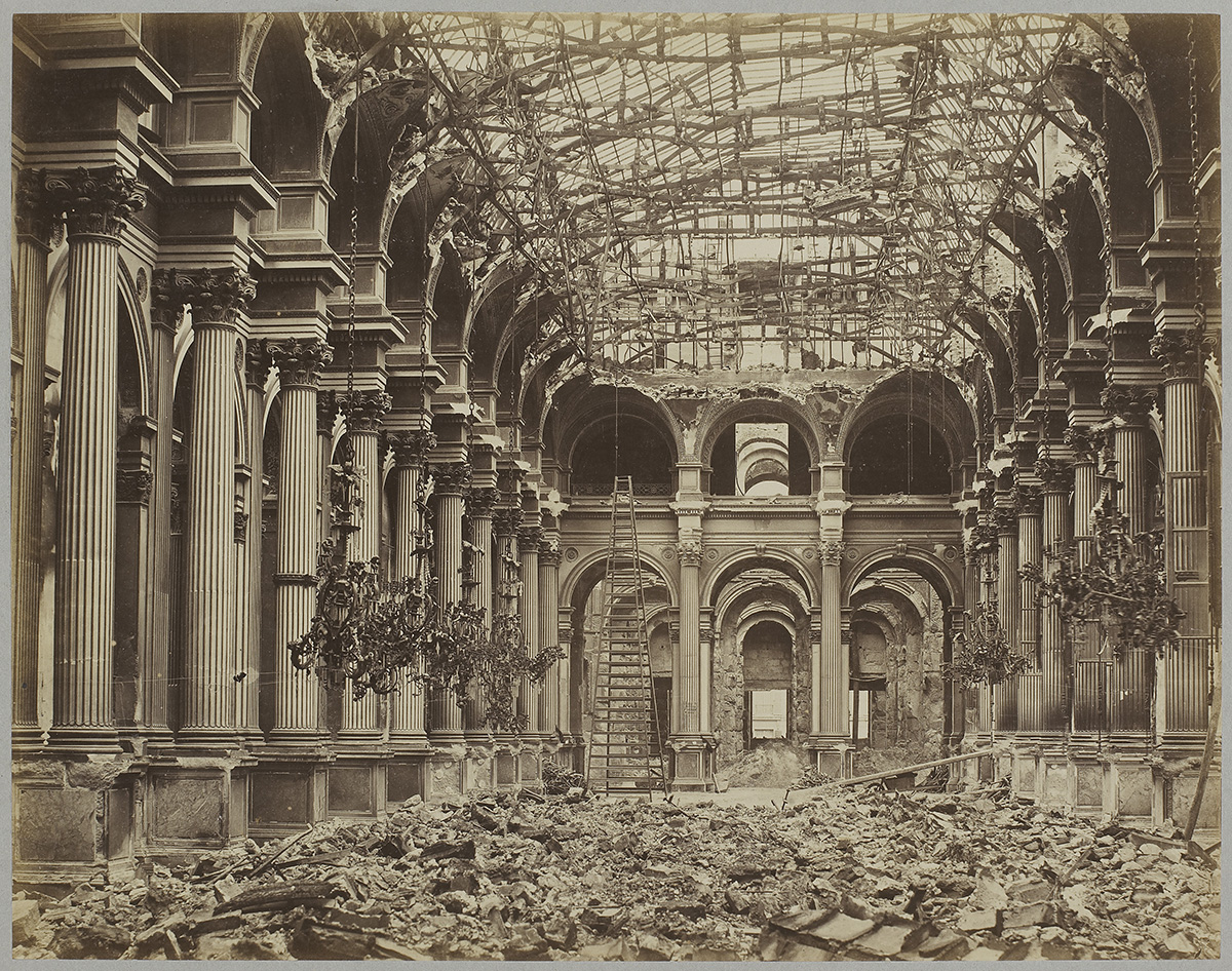 Black-and-white photograph of a ruined interior. Fluted stone columns support a ceiling of mangled iron beams and melted chandeliers while debris covers the floor.