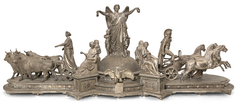 Metallic sculpture with a winged and draped female figure standing on a dome. She holds wreaths in her outstretched hands. Seated figures and cherubs surround her; an eagle spreads its wings below, at center. Standing figures tend animals on either side—oxen on one side and horses with a chariot on the other.