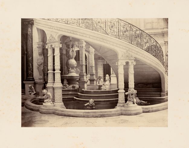 Black-and-white photograph of a stepped fountain with stone cherubs playing in water beneath a curved staircase supported by ornamented, paired columns.