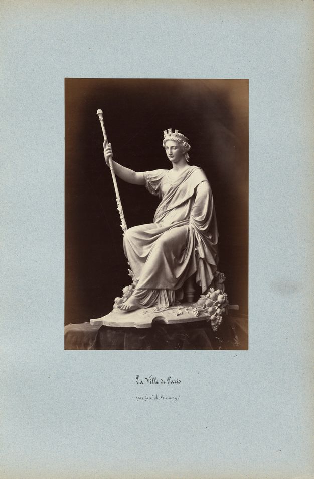 Black-and-white photograph on a blue page showing a model for a seated and draped female figure holding a staff. The handwritten caption below the photograph indicates that the sculptor is deceased.