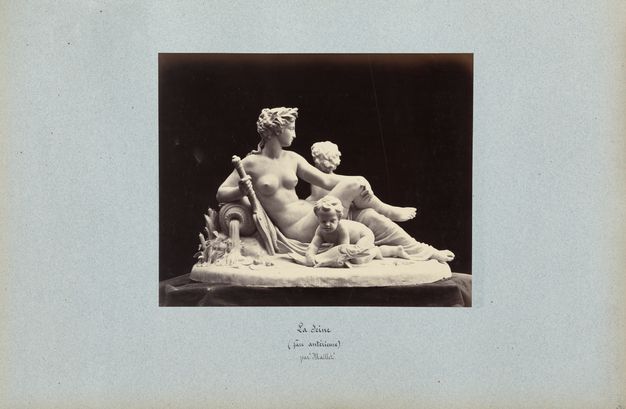 Black-and-white photograph on a blue page showing a model of a nude woman reclining against a pouring vase while holding an oar. A cupid in the foreground grasps a fish. A handwritten caption is below.