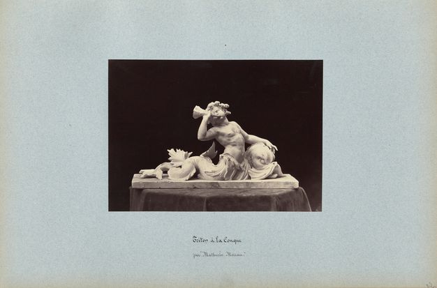 Black-and-white photograph on a blue page showing a model for a twisting nude male figure whose lower torso becomes a fish tail. He blows into a shell held in one hand. A handwritten caption is below.