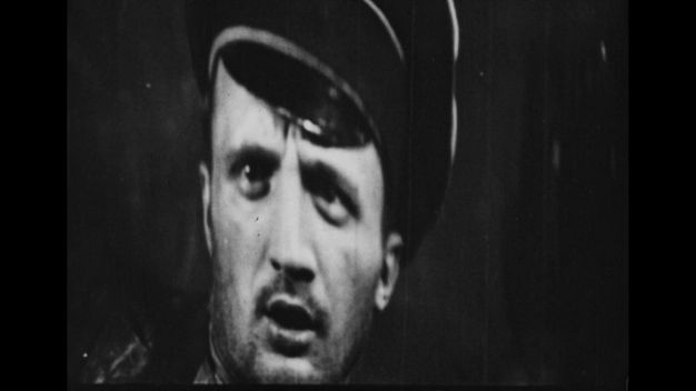 The second of three black-and-white still frames shows the face of a man in a uniform cap staring slightly off camera in shock, with his mouth open.