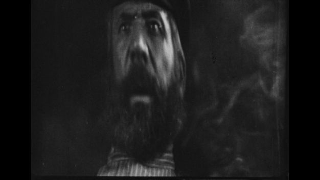 The third of three black-and-white still frames shows a thickly bearded man with parted lips staring beyond the viewer in horror.