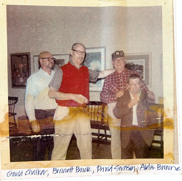 A creased and slightly discolored photo showing four men of middle age or older standing in a room with paintings on the wall behind them, against which are a few wood chairs. Buck wears glasses and a red vest, holds a cigarette, and affectionately puts his arm on another man’s shoulder. Their names are handwritten in blue pen below.