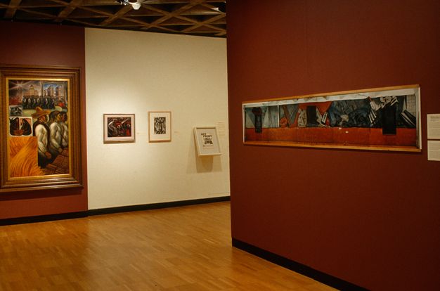A photograph of a museum gallery with wood floors. On a red wall at right is a framed horizontal reproduction. On a back wall hangs figure 1, in a heavy frame, adjacent to three smaller works that are barely visible.