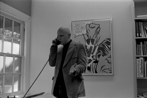 A black-and-white photograph of a man in a sport coat standing, talking on the phone, and gesticulating; his gaze turns downward. Behind him is a framed print and a bookshelf.