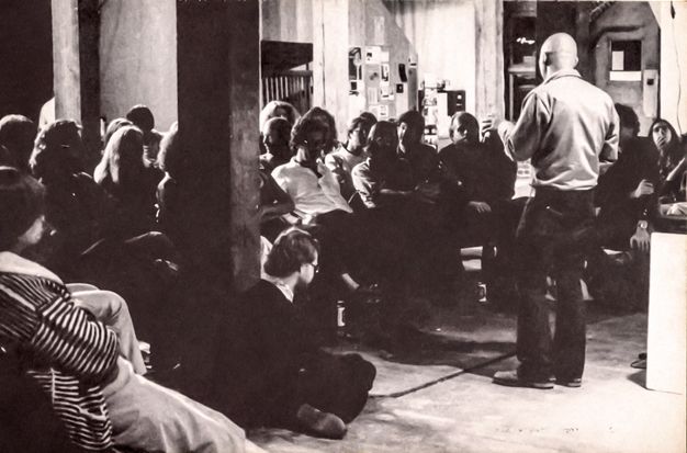 A black-and-white photograph of a man viewed from behind as he stands, delivering a talk to a seated, intimate audience in a full room. Two wooden pillars bisect the room, and one audience member sits cross-legged on the floor.