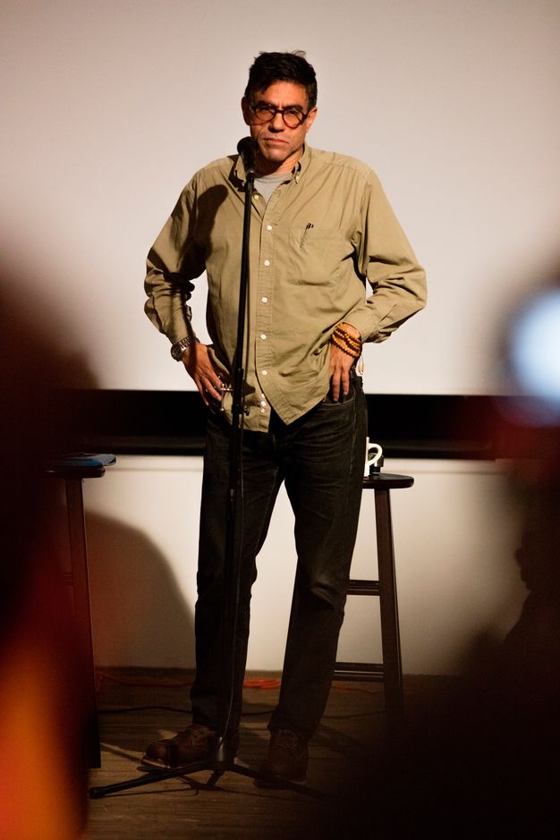 Color photograph of a man in glasses standing in a casual button-down khaki shirt with his hands on his hips in front of a stool, gazing at the viewer and an unpictured audience.