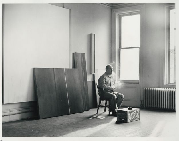 A black-and-white photograph of a man seated on a chair smoking a cigarette inside a window-lit room, which is bare except for the artworks stacked against the wall behind him and an upside-down Pepsi-Cola crate on the hardwood floor.