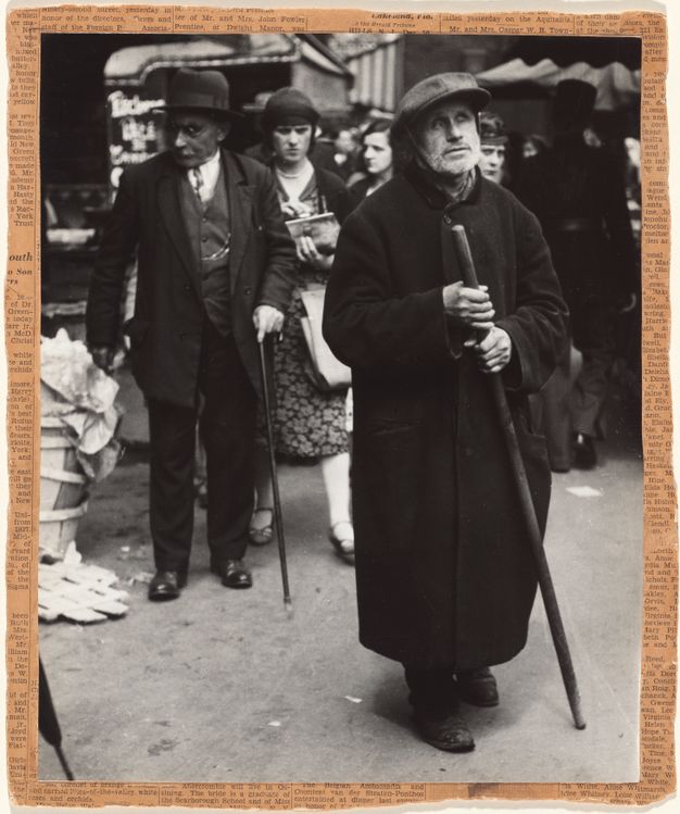 A black-and-white photograph of a white-bearded man wearing a long coat and flat cap; he looks up as he walks with a long stick in a crowded street. A man and woman walk behind him. A yellowing newspaper with uneven edges borders the image.