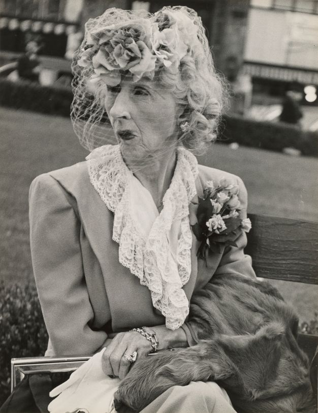 A black-and-white photograph of an older woman sitting on a bench wearing a floral cocktail hat with white-netted veil, a blazer with laced ruffles, jewelry, and a boutonniere. A fur coat is draped over her arm. Her lips are pursed pensively.