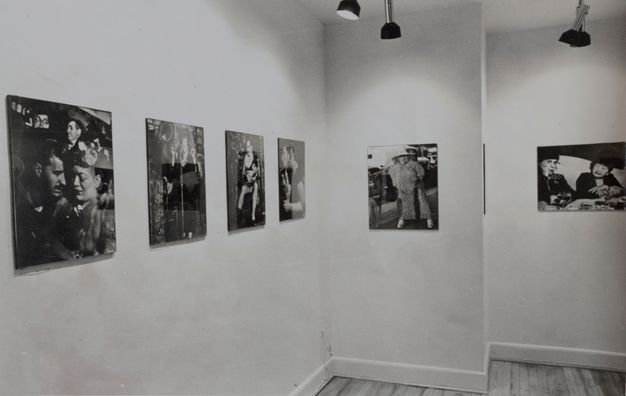 A black-and-white photograph showing seven black-and-white photographic prints of people hung at eye-level in a gallery; the installation wraps two small corners within the space.