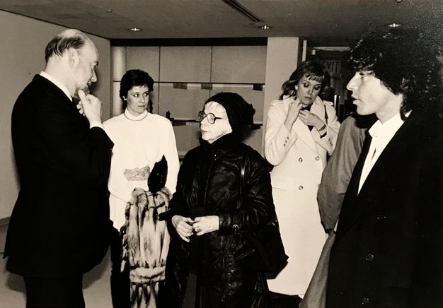 A black-and-white candid photograph of six well-dressed individuals who engage in separate activities in an interior space. A man on the viewer’s right observes the conversation between the taller Lunn, who traces his beard, and Model, who looks down; a woman in the background holds her patterned coat and looks askance toward another woman, who touches her own scarf.
