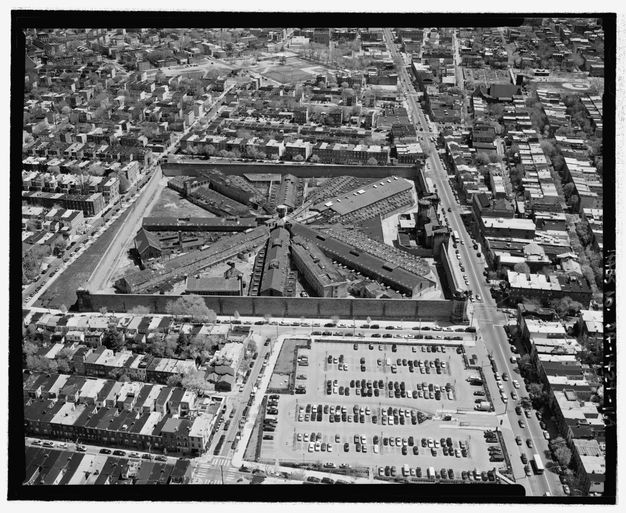Black-and-white photograph showing the radial plan of the penitentiary’s buildings, rectilinear perimeter wall, and surrounding areas, including a parking lot, a park, streets with cars, and blocks of urban housing.