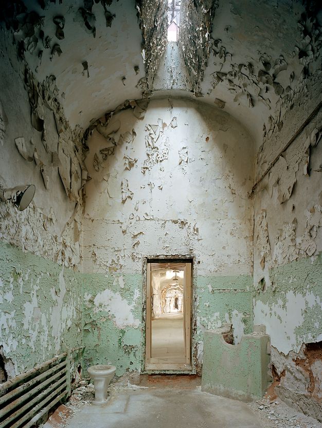 Color photograph of the view from inside a deteriorated, unfurnished cell with green and white peeling walls toward the open doorway, which shows a shimmering reflection of the external corridor through a mirror positioned in the corridor between cell doors and turned diagonally.