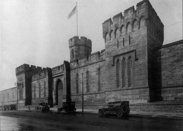Black-and-white photograph of a street view of the original Gothic Revival facade with crenellated towers; Model-T cars are parallel parked in front of the gate.