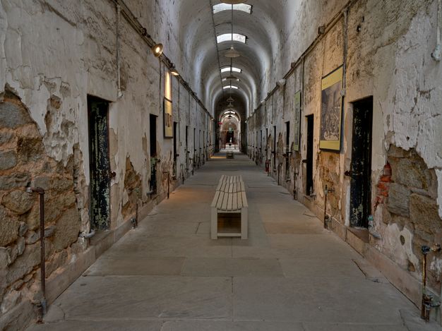 Color photograph of a view down a long corridor toward a center courtyard; cell doorways on either side are set into deteriorating stone walls, on which museum signage is hung; and skylights at the top of the barrel-vault ceiling bring some light into the space.